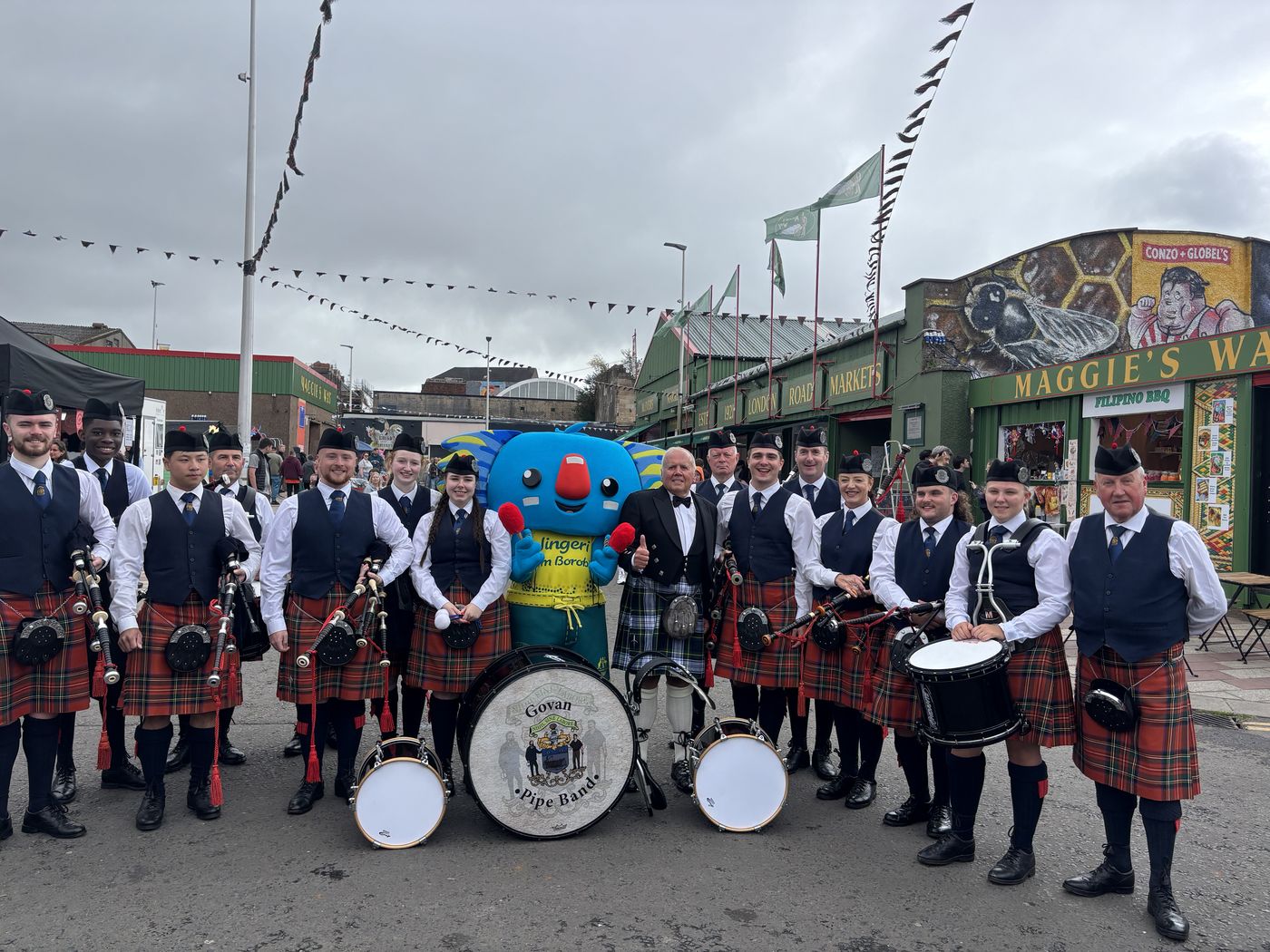 The full Govan Pipe Band lined up around their bass drum on a city street, with a blue mascot in the middle