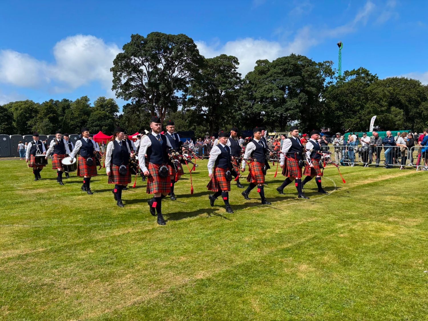 Govan Pipe Band pipers and drummers marching across a field in red tartan kilts and navy waistcoats